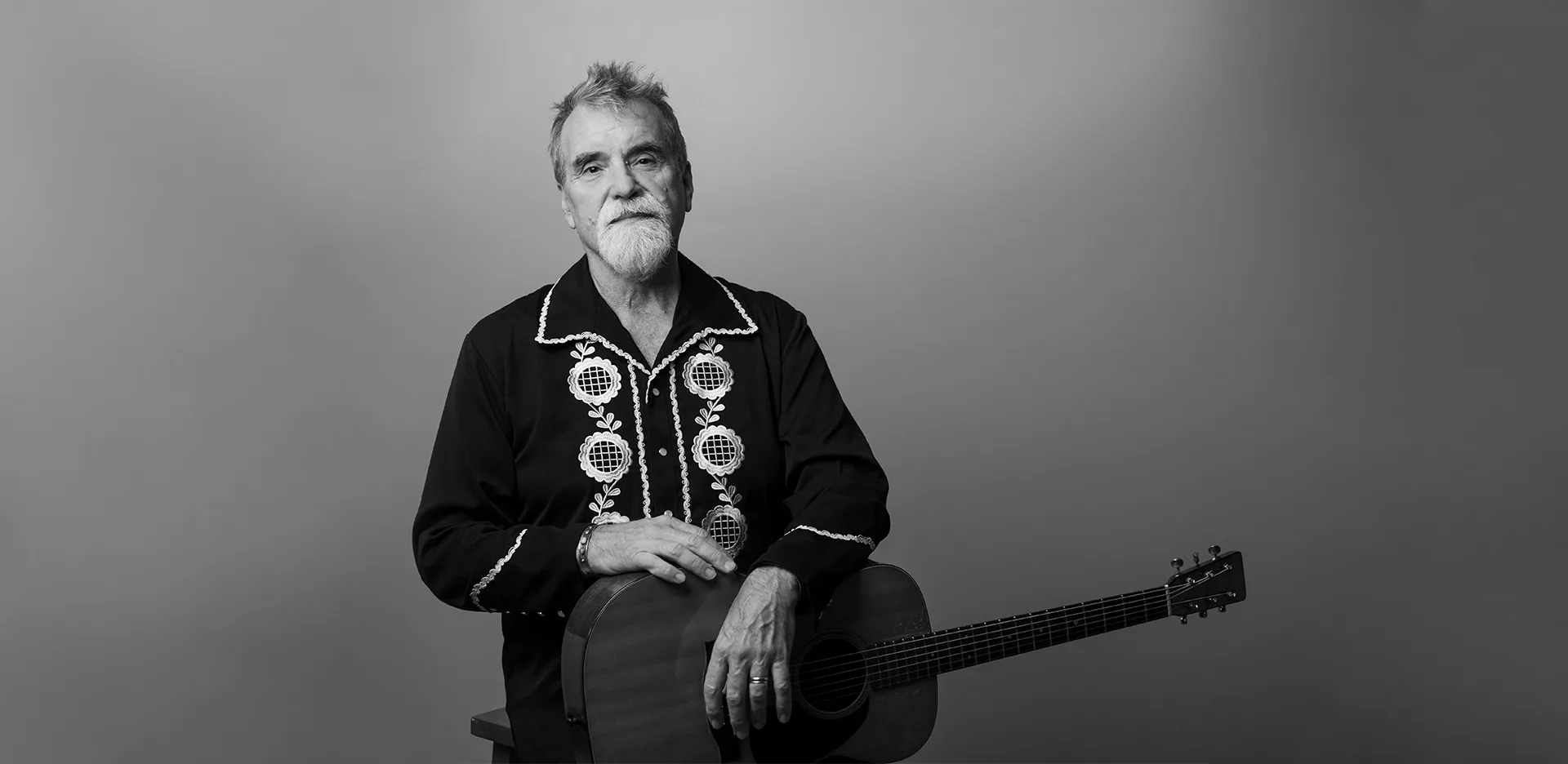A grayscale photo of an older man with short hair and a beard, wearing a decorated dark shirt, standing with his hands resting on an acoustic guitar against a plain background.