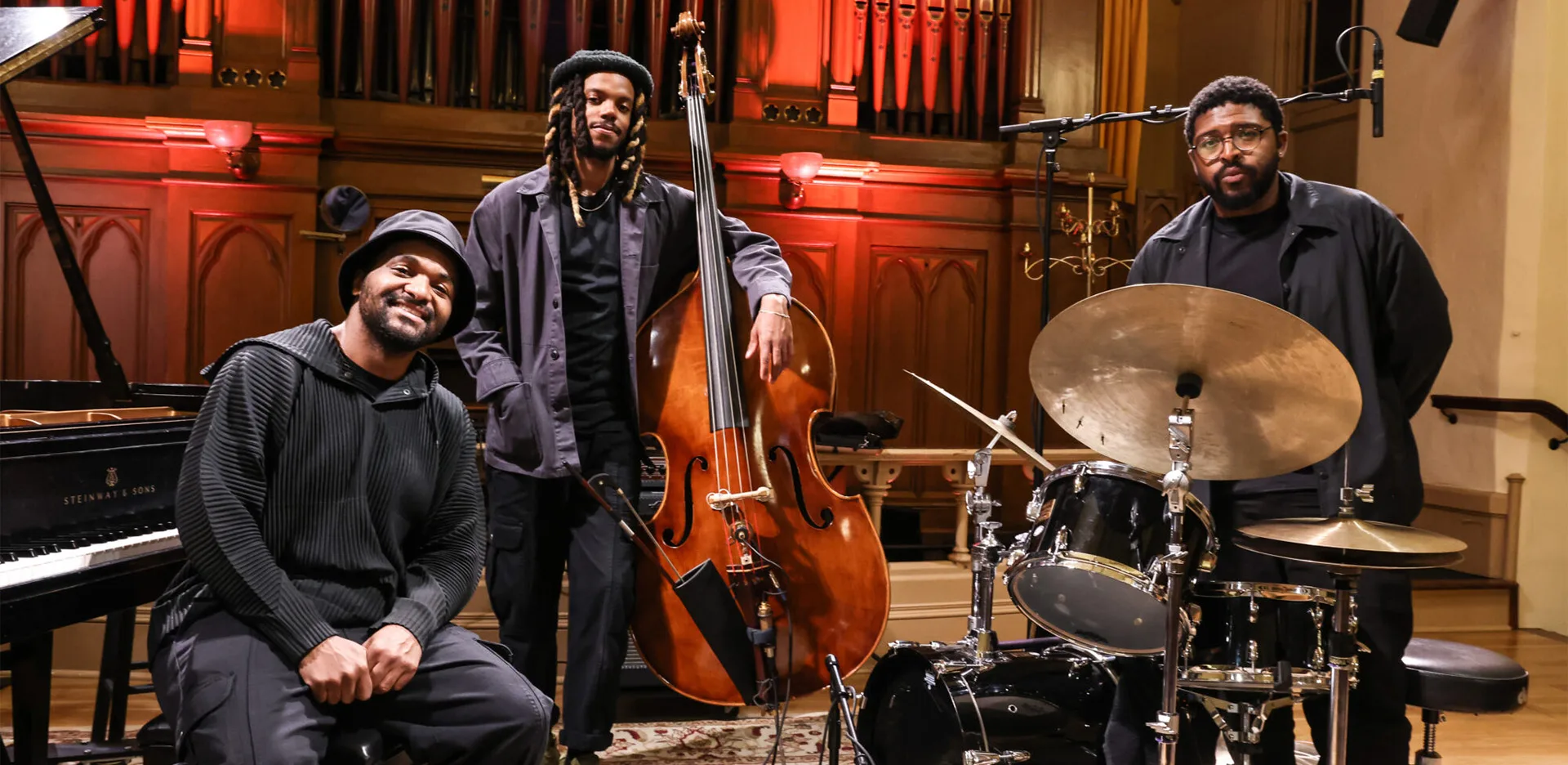 Three musicians pose in a warmly lit concert hall with wooden organ pipes in the background. One sits at a piano, another stands with a double bass, and the third stands behind a drum set. All are dressed in dark clothing.