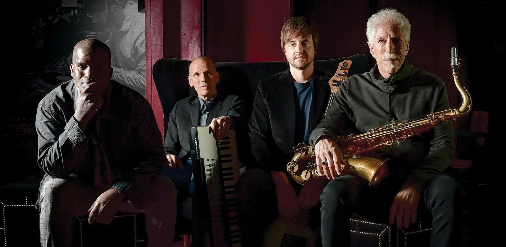 Four men sit indoors with musical instruments; one holds a saxophone, another a keyboard, one rests his hand on a guitar, and the fourth sits pensively. The background is dark with hints of deep red tones.