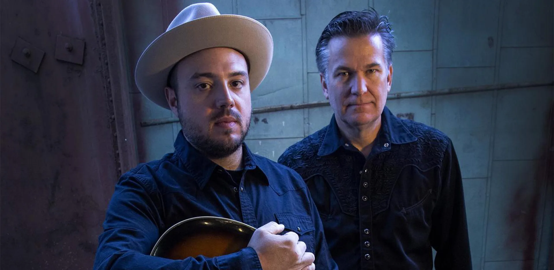 Two men stand side by side, both wearing dark shirts. The man on the left wears a light hat and holds a guitar, while the man on the right stands slightly behind him. The background is industrial and blue-toned.