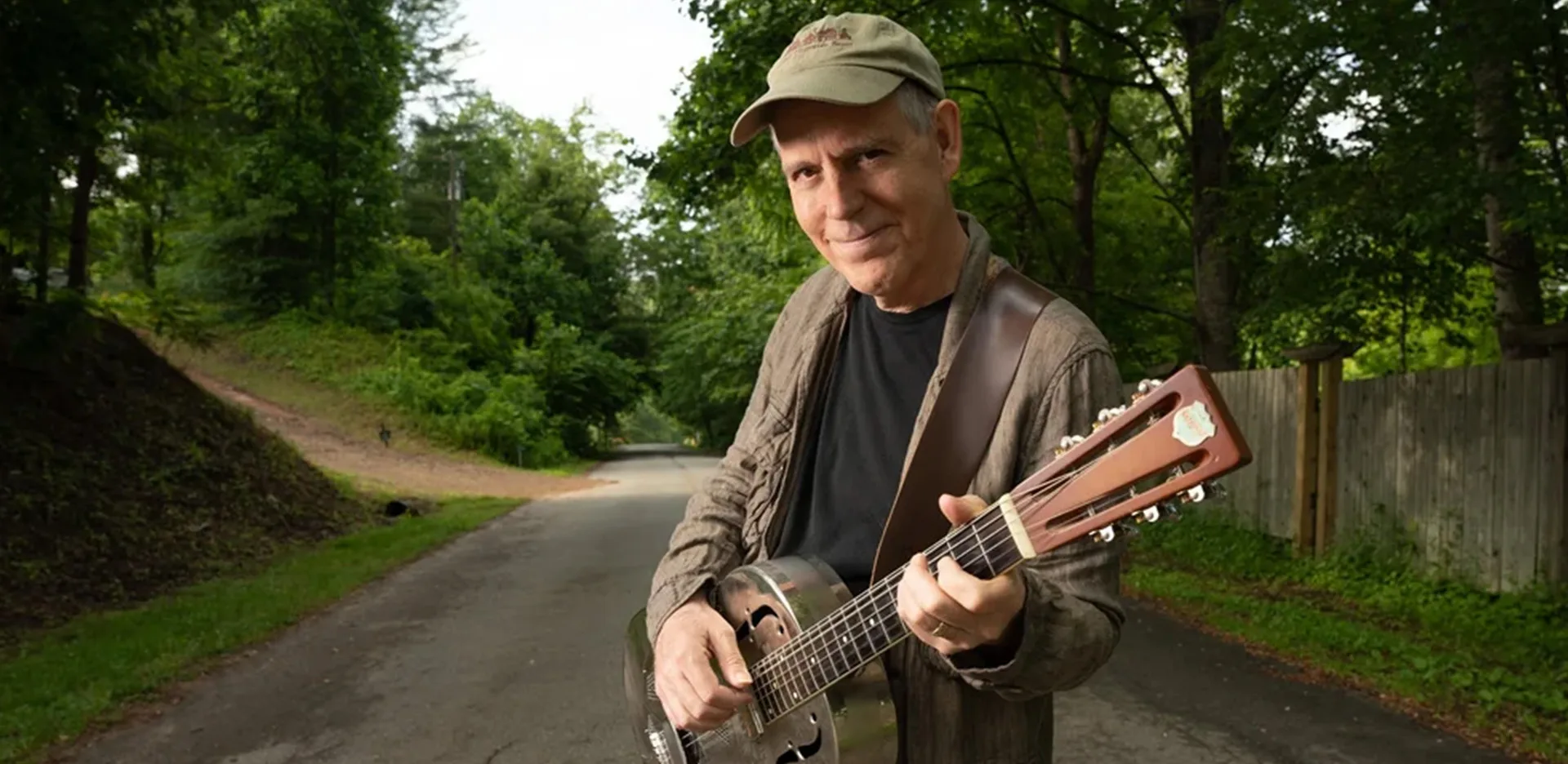A man stands on a quiet, tree-lined road playing a resonator guitar. He wears a cap, a jacket, and a black shirt, smiling warmly at the camera. The background is lush and green with a wooden fence on the right.