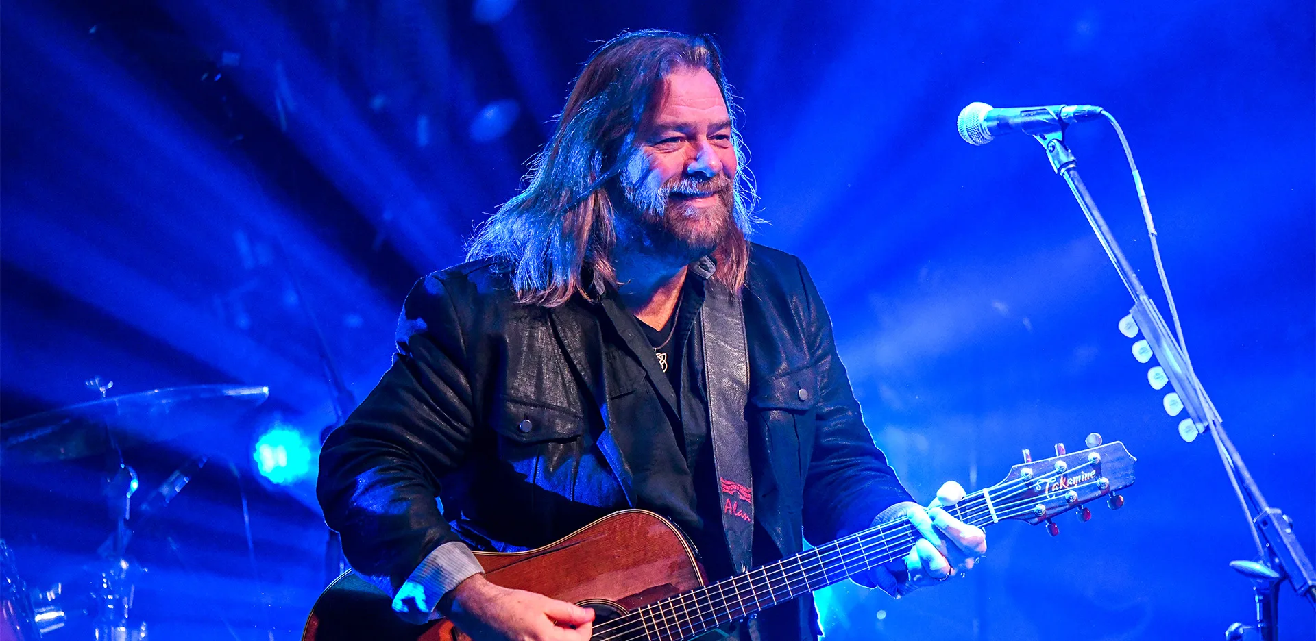 A man with long hair and a beard plays an acoustic guitar and smiles on stage, illuminated by blue lights, with a microphone stand in front of him.