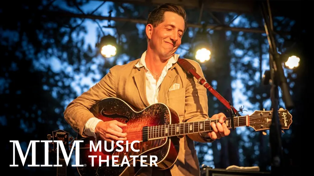 A smiling man in a tan suit plays an acoustic guitar on an outdoor stage with bright lights. The background shows trees at dusk. The text “MIM Music Theater” appears in the lower left corner.
