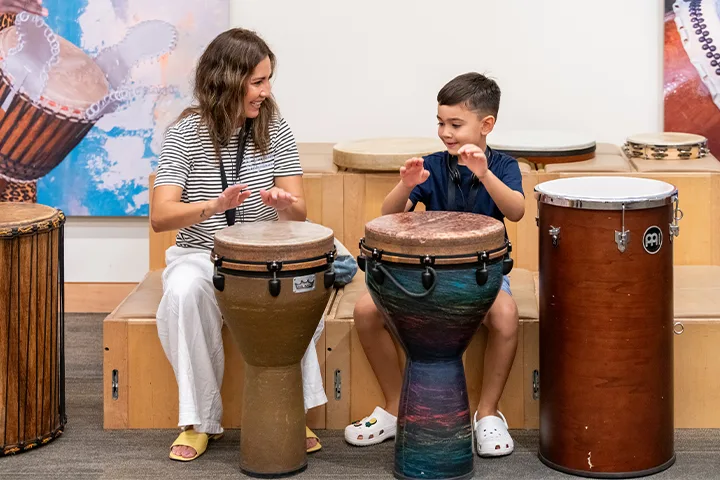 A woman and a young boy sit side by side playing different hand drums, smiling and looking at each other in a bright room with several drums in the background.