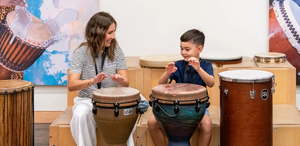 A woman and a young boy sit side by side playing hand drums, smiling at each other. Colorful drums and drum artwork are visible in the background, creating a lively, musical atmosphere.