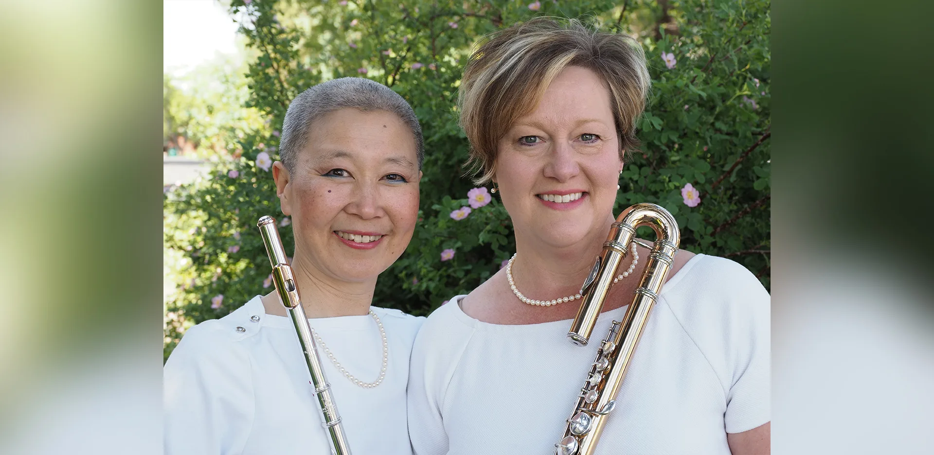 Two women smiling outdoors, holding flutes. Both wear white tops and pearl necklaces, standing in front of green foliage with pink flowers.