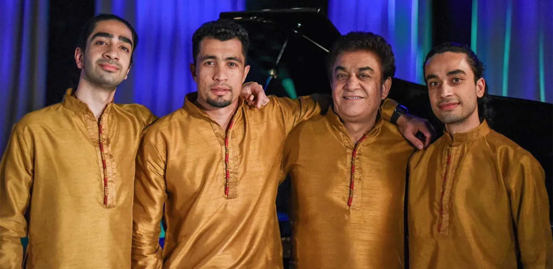 Four men wearing matching golden traditional outfits stand side by side, smiling, with arms around each other. A piano and blue curtains are visible in the background.