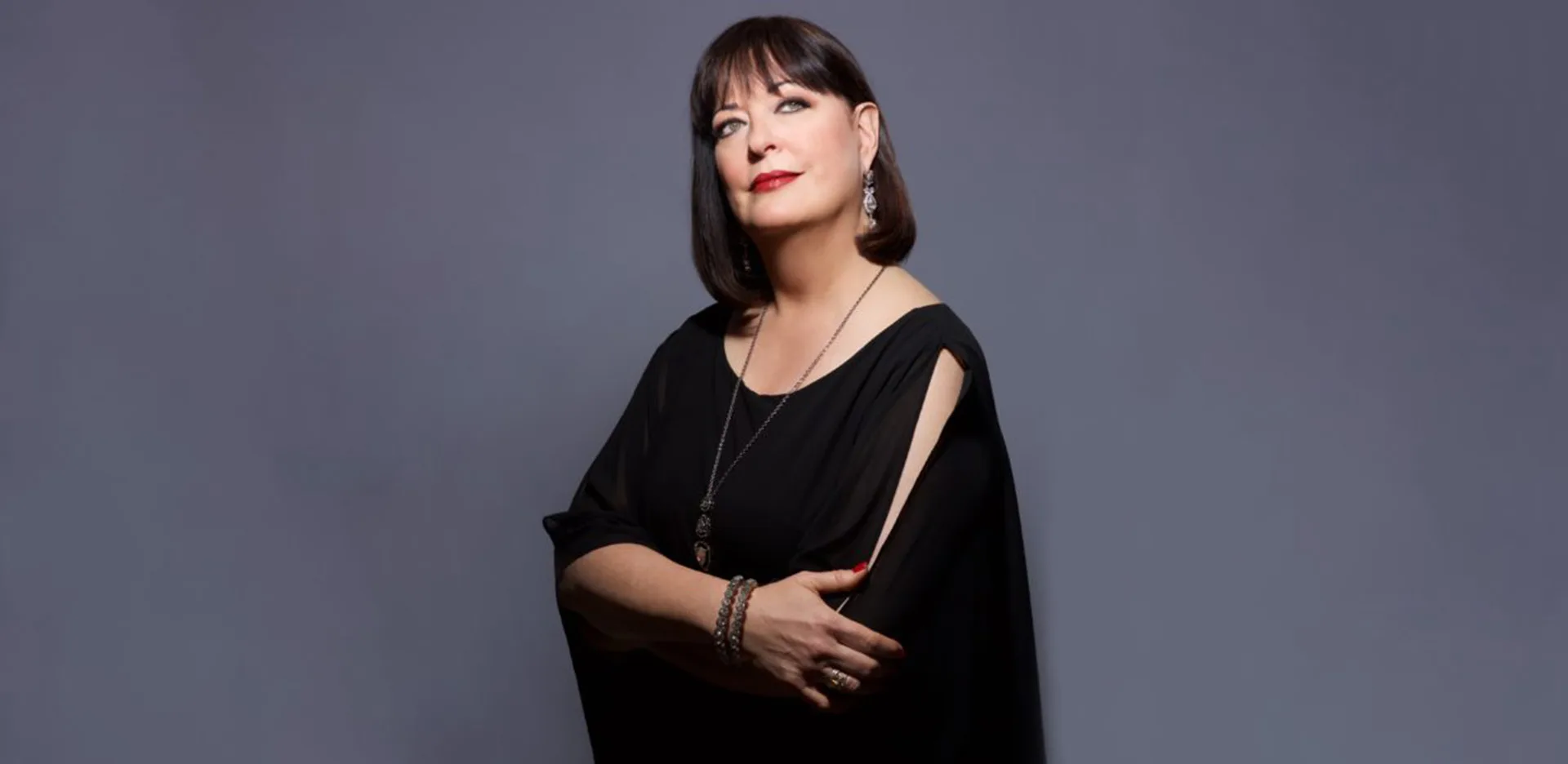 A woman with straight dark hair and bangs, wearing a black dress and jewelry, stands against a plain gray background with her arms crossed and a thoughtful expression.
