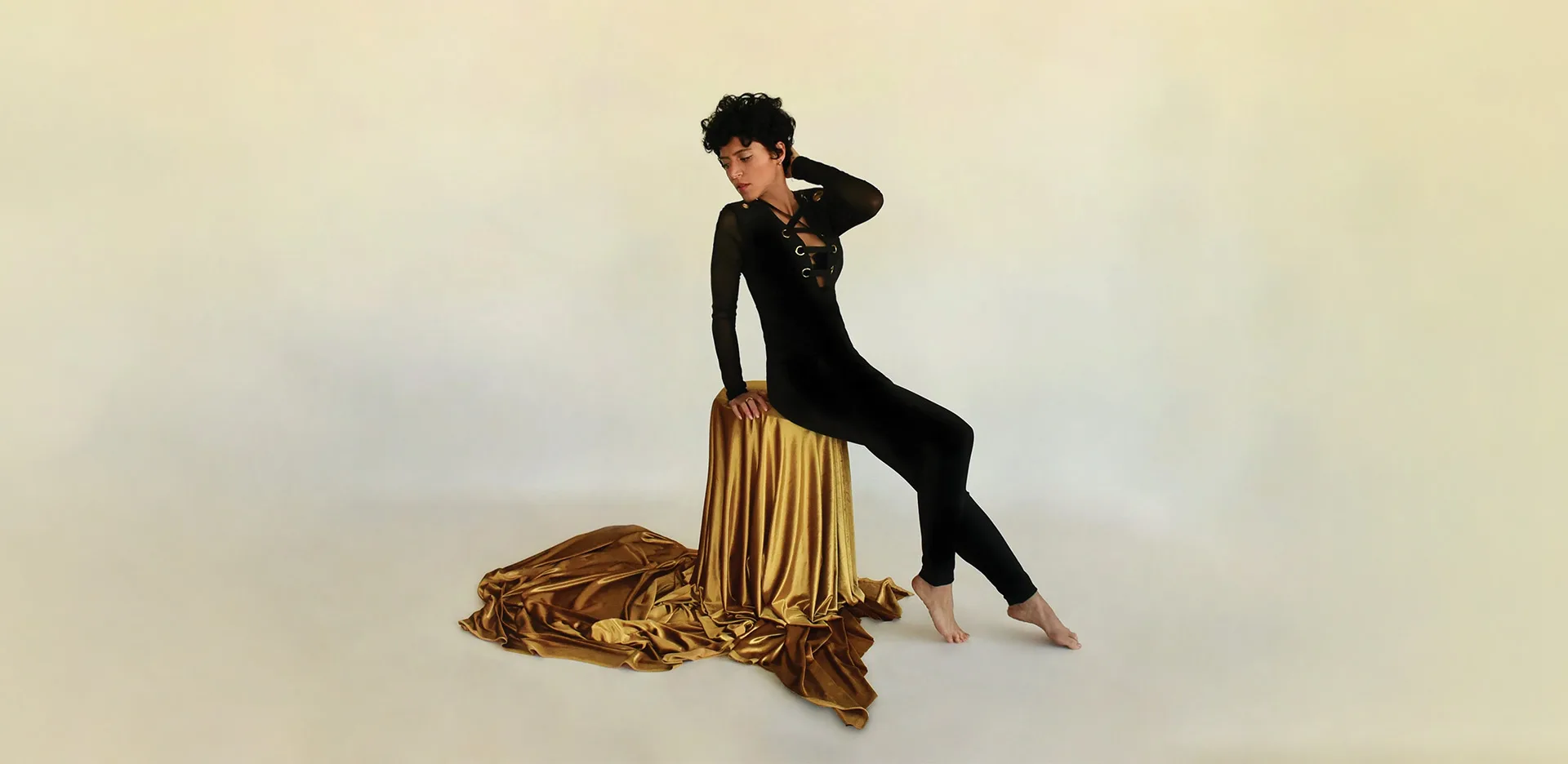 A person in a fitted black outfit poses barefoot on a gold-draped stool against a plain light background, with gold fabric pooled on the floor.