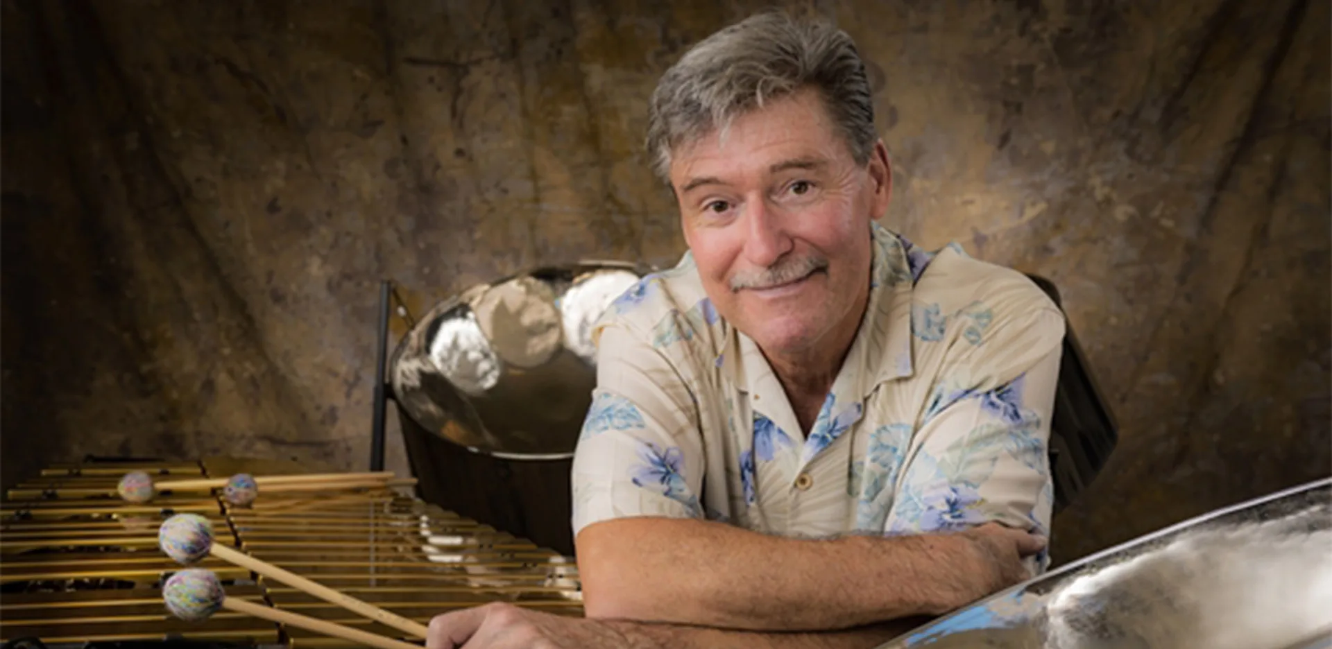 A smiling older man in a floral shirt leans on a marimba with colorful mallets, with steel drums in the background and a brown textured backdrop behind him.
