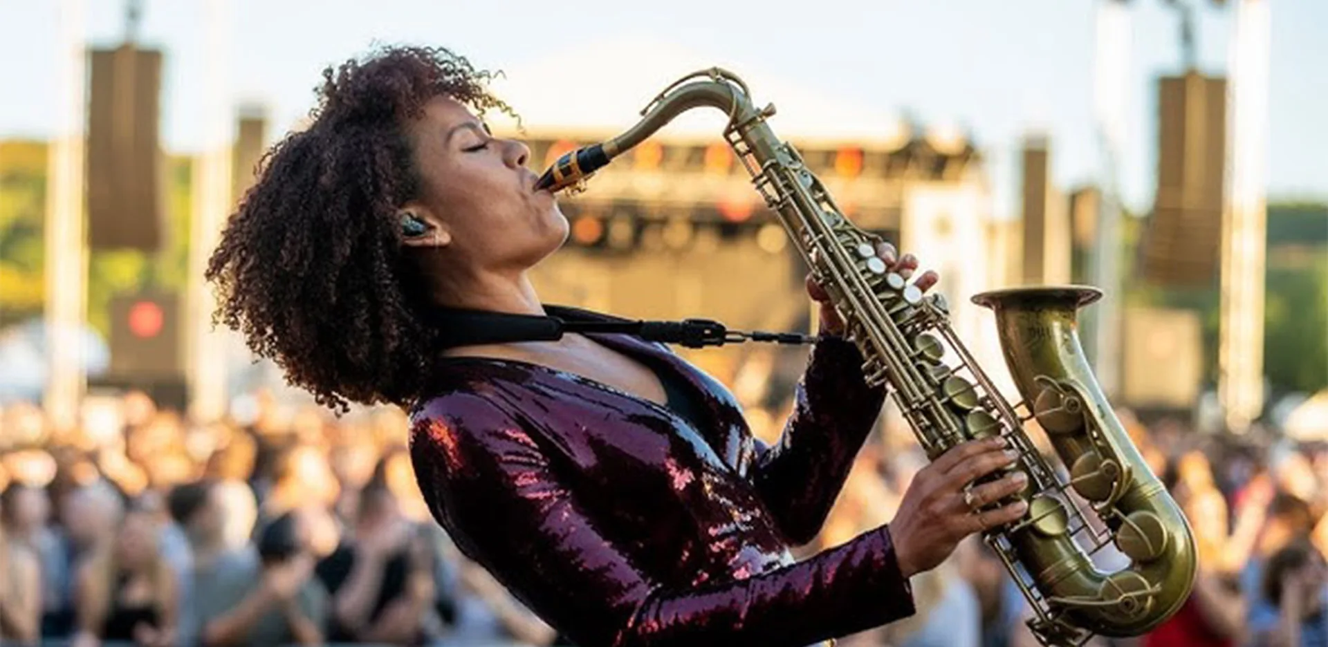 A woman wearing a shiny, burgundy jacket plays a saxophone passionately on an outdoor stage, with a blurred crowd and concert equipment in the background.