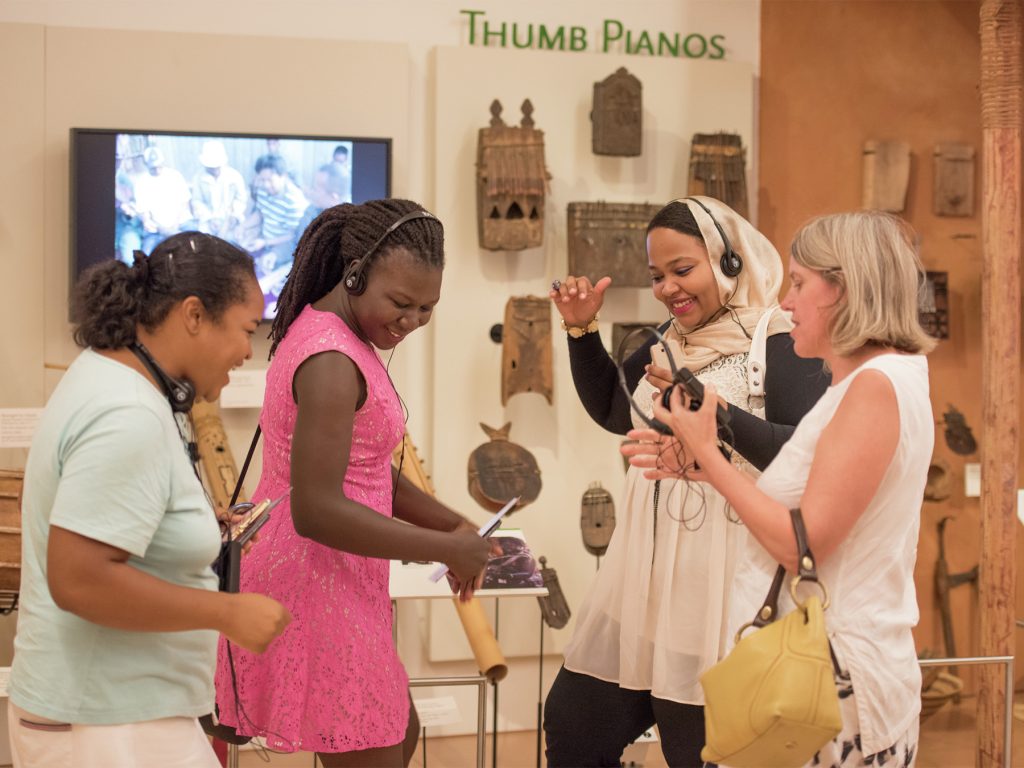 Four guests wearing Guideport headsets dance together in front of the Thumb Pianos exhibit.